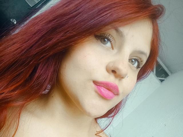 Foto em close-up de Sophie Hill, com cabelos ruivos longos, pele clara e batom rosa. Ela está olhando diretamente para a câmera com um leve sorriso, transmitindo uma vibe sexy e sedutora. Iluminação brilhante.