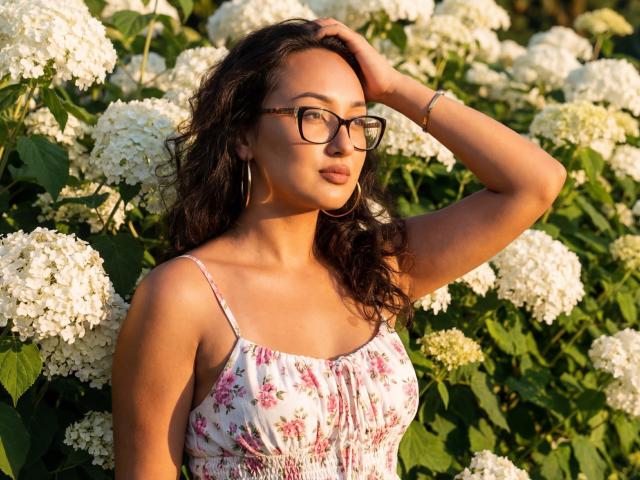 EvaSarazardje, uma mulher atraente de cabelos escuros e óculos, vestindo um vestido floral, com a mão no cabelo, em frente a um cenário exuberante. Hortênsias brancas. A luz do sol realça sua pele bronzeada e sua postura confiante. Absolutamente deslumbrante..