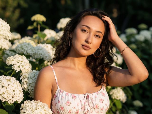 EvaSarazardje, uma mulher asiática atraente com cabelos pretos ondulados, vestindo um vestido branco com estampas florais, com a mão no cabelo, cercada por branco. flores. A luz do sol realça sua pele bronzeada e seu olhar confiante. Transmite uma vibe sedutora..