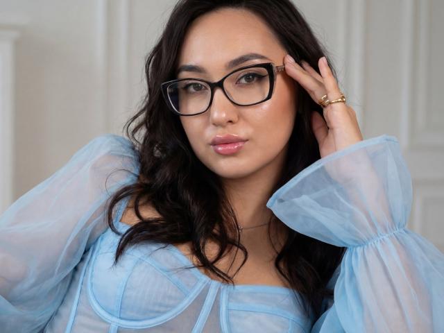 EvaSarazardje a de longs cheveux noirs, porte des lunettes noires et une robe bleue transparente avec des manches bouffantes. Elle touche ses cheveux avec sa main droite, tout en portant une bague en or. Un look incroyablement séduisant..