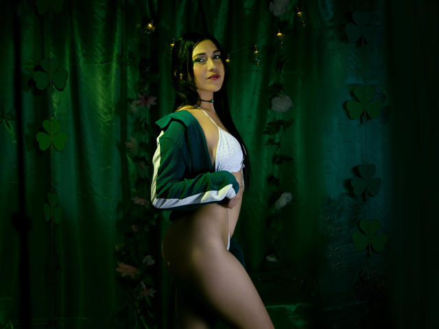 Photographie de Valentina Moreau, avec de longs cheveux noirs, portant une veste noire par-dessus un soutien-gorge blanc, debout de côté, montrant son corps voluptueux et sexy, ses cuisses nues, un fond floral sombre. Extrêmement sexy.