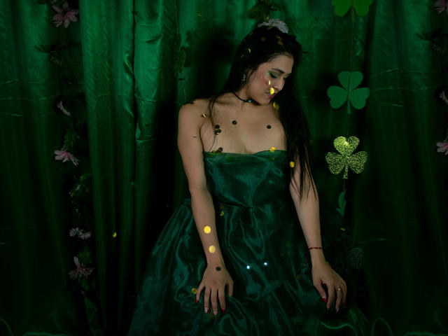 Valentina Moreau, con i suoi lunghi capelli neri, indossa un abito scuro verde senza spalline, illuminata da una luce gialla e verde brillante. luci. È in piedi di fronte a tende verde scuro con motivi floreali. Assolutamente attraente..