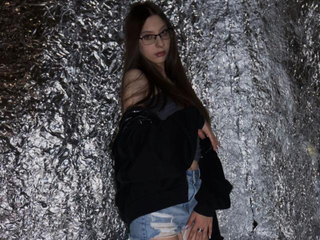 RavenBlisss, eine junge Frau mit langen, dunklen Haaren und einer Brille, lehnt sich an eine glänzende, metallisch strukturierte Wand. Sie trägt ein schwarzes Oberteil und abgenutzte Jeans und wirkt dabei verführerisch. Verdammt heiß..