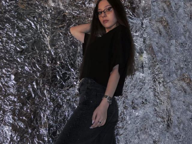 RavenBlisss está apoiada em um fundo prateado texturizado e enrugado. Ela veste uma blusa e uma saia pretas, óculos de sol escuros e um relógio. A mão dela repousa sobre a cabeça, transmitindo uma aura confiante e sensual..