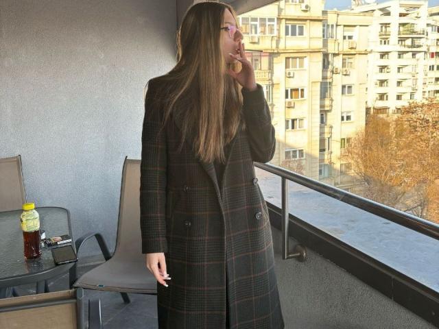 RavenBlisss está de pie en un balcón, vistiendo un abrigo largo de cuadros negros y fumando un cigarrillo. Ella está mirando hacia los altos edificios de apartamentos, con una silla y una botella cerca. Hojas de otoño en el fondo.