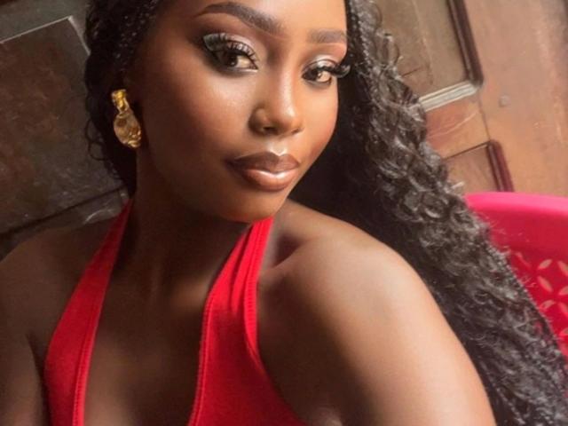 Gros plan sur SexyDivanah-hot, une femme noire attrayante avec de longs cheveux bouclés, portant un haut rouge à encolure américaine, des boucles d'oreilles dorées. et un maquillage audacieux. Ses lèvres pulpeuses et ses yeux intenses dégagent une tension sexuelle..