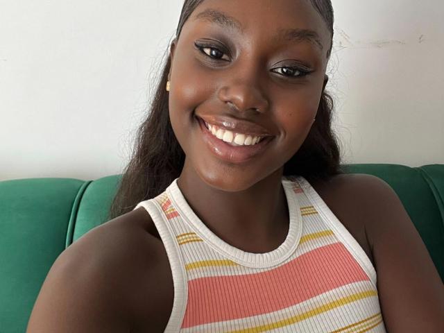 Close-up de SexyDivanah, muito atraente, com pele escura, sorrindo amplamente, mostrando dentes brancos, vestindo uma camiseta branca com detalhes vermelhos e listras amarelas, sentada em um sofá verde. Luz natural e abundante.