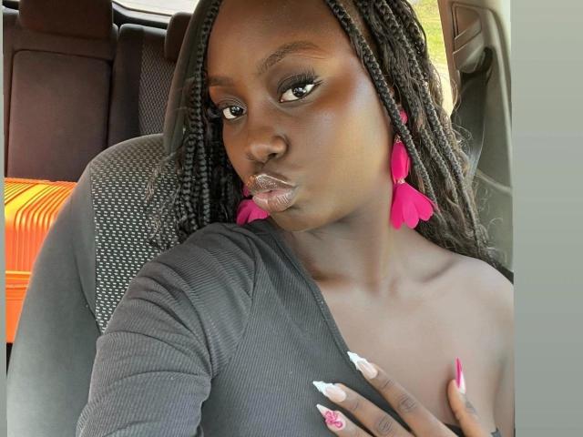 SexyDivanah - atraente, de pele escura, com longas tranças, usando brincos de franjas rosa, blusa preta e unhas brancas, com os lábios franzidos. lábios, mão no peito, assento de carro com tecido laranja ao fundo, olhar intenso..