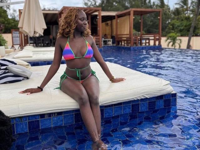 SexyDivanah - aantrekkelijk, donkerhuidig met krullend rood haar, zittend op de rand van het zwembad, gekleed in een kleurrijke bikini, benen gekruist. zwembad met blauwe tegels, houten hutjes op de achtergrond, sensuele sfeer, waterdruppels op de huid.