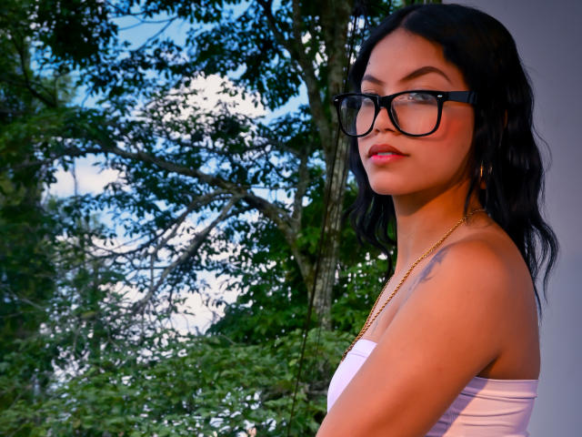 Photograph of MaliaFoxxy, standing outdoors, side profile, wearing black-framed glasses, white strapless top, gold necklace, with long black hair, intense gaze, sunlight highlighting her face, trees in background. Fucking sexy.