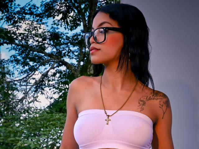 Photograph of MaliaFoxxy, with black hair, wearing black glasses and a white strapless top, gold necklace, and tattoo on her right shoulder. She gazes to the side with a sultry expression.