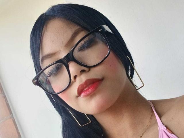 Primer plano de MaliaFoxxy, una joven con cabello largo y negro, que lleva gafas negras, lápiz labial rojo y una blusa rosa. Ella está mirando directamente a la cámara con una sonrisa ligeramente seductora..