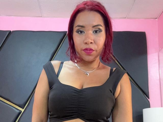 Fotografia de LinaBowie com cabelo ruivo na altura dos ombros, vestindo uma blusa preta justa que realça seu corpo de tamanho médio. seios, e um colar de prata. Ela tem batom rosa e uma expressão neutra. Incrivelmente atraente..