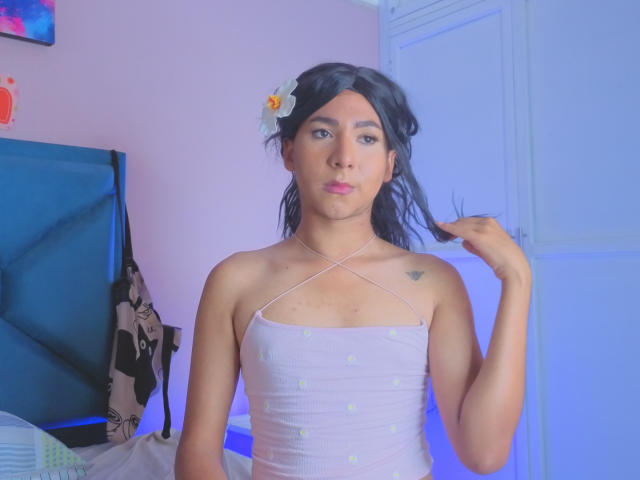 ZoeHotMoretti, stehend in einem Schlafzimmer, trägt ein weißes, trägerloses Top mit Halstuch und ein Kleid, schwarzes Haar, eine Blume im Haar, hält... Haare, blaues Licht, schwarzer Kopfteil, Stoff mit Blumenmuster, ernster Gesichtsausdruck.