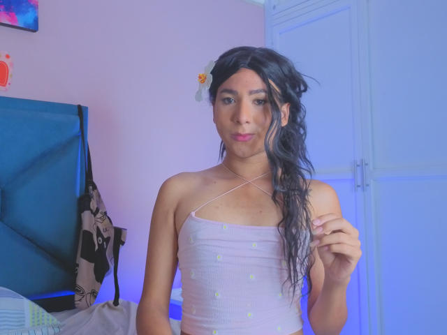 ZoeHotMoretti stands in a room, wearing a white, strapless top with small buttons, and a white flower in her wavy, black hair. She holds a necklace, blue lighting in background.