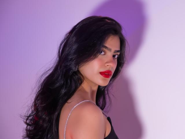 Fotografía de Ruby Carson con cabello negro largo, lápiz labial rojo y un vestido negro, mirando por encima del hombro. Mirada seductora, resaltada por una iluminación púrpura y blanca. Increíblemente atractivo/a..