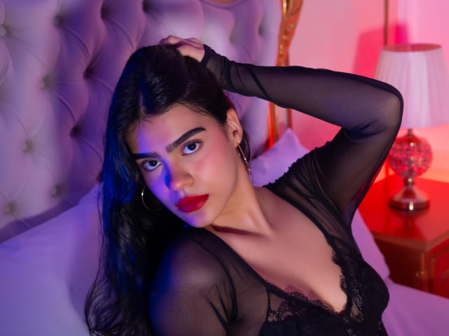 RubyCarson, dark-haired woman with red lipstick, black lace top, hand in hair, seductive gaze, tufted headboard, red and blue lighting, bedside table with lamp, sexual tension, intimate setting.
