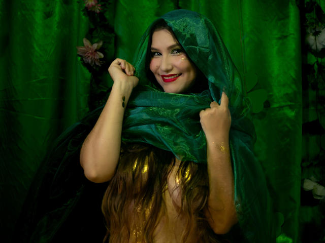 Maria Rowe è al centro, con un mantello verde con cappuccio che copre parzialmente le sue spalle e il suo seno. Ha il rossetto rosso, gli occhi gialli luminosi e i capelli lunghi castani. Sfondo verde scuro. Incredibilmente sexy..