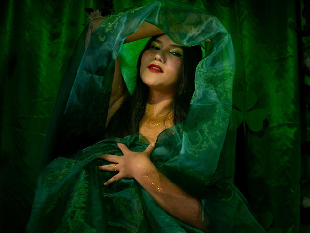 Maria Rowe, vêtue d'un voile transparent vert foncé, sa main agrippant le tissu, les yeux à moitié clos, les lèvres rouges entrouvertes, des paillettes dorées sur... sa peau, une forte tension sexuelle, une palette de couleurs vert et noir.