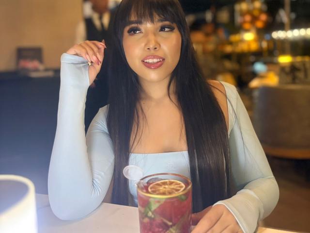 Heyly, avec ses longs cheveux noirs et sa frange, portant un haut bleu clair à une épaule, assise au bar, tenant un verre. plan rapproché, aspect séduisant, arrière-plan flou, éclairage vif.