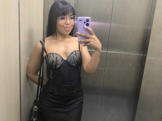 Heyly with straight black hair, glasses, and a black strapless dress with sparkly details, takes a mirror selfie in an elevator. She's smiling, holding a phone with three cameras.