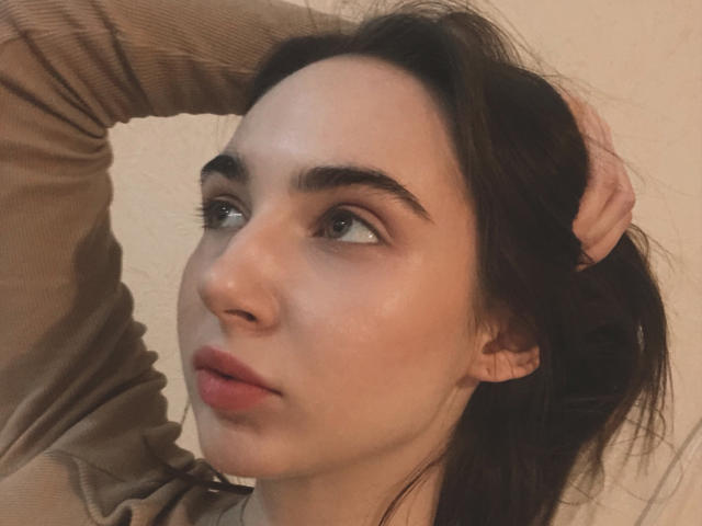 Gros plan de Miiraii, une jeune femme à la peau pâle, aux cheveux bruns foncés et aux sourcils épais. Elle regarde vers le haut, la bouche légèrement ouverte, une main dans ses cheveux, et porte un haut marron. Extrêmement séduisant..