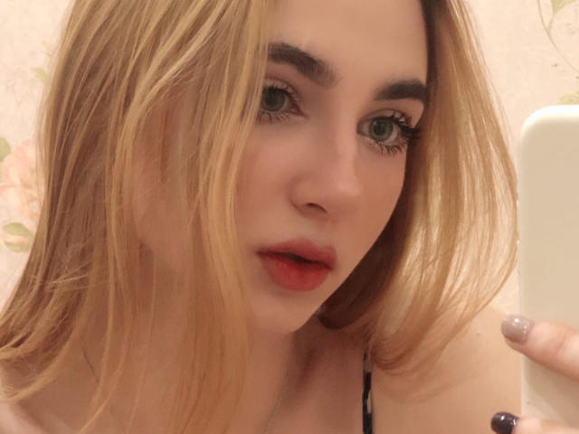 Autorretrato de Miiraii en primer plano, cabello rubio que cubre parcialmente el rostro, labios rojos, ojos azules intensos, labios ligeramente entreabiertos. piel clara, correa negra en el hombro, mano derecha tocándose el pelo, tensión sexual..