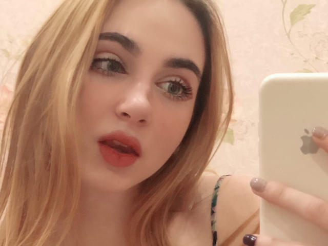 Selfie de primer plano de Miiraii con cabello largo y rubio, lápiz labial rojo y cejas oscuras, sosteniendo un iPhone con el logo de Apple. logotipo. Sus ojos están medio abiertos, lo que le da una mirada seductora. Las uñas están pintadas de un morado oscuro..