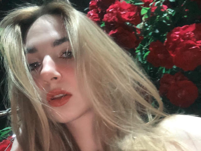 Nahaufnahme von Miiraii mit blonden Haaren, roten Lippen und einem verführerischen Ausdruck, umgeben von leuchtend roten Rosen. Das weiche Licht betont ihr Gesicht und erzeugt eine verführerische, intensive Atmosphäre..