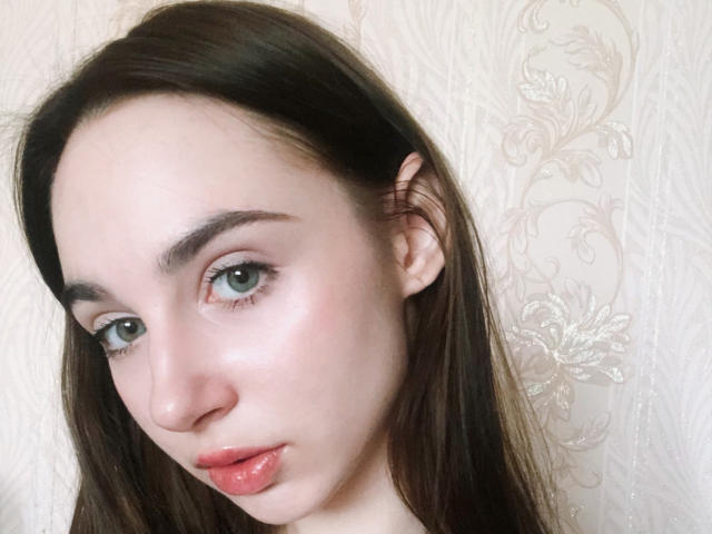 Fotografía de primer plano de Miiraii, una mujer de piel clara con cabello largo y castaño oscuro, ojos azules y labios rojos brillantes, que está mirando. Mirando directamente a la cámara con la cabeza ligeramente inclinada. Fondo blanco con textura..