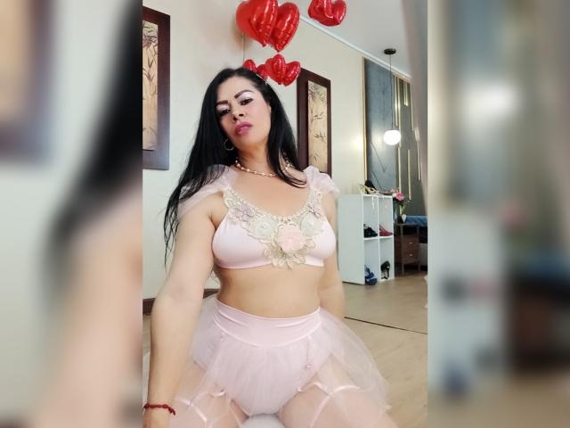 SofiMontes steht in einem Schlafzimmer und trägt einen durchsichtigen, rosa Spitzen-BH und einen Tutu, wobei ihre großen Brüste und ihre kurvigen Hüften sichtbar sind. Rote Herzballons schweben über ihrem Kopf und erzeugen eine sinnliche Atmosphäre..