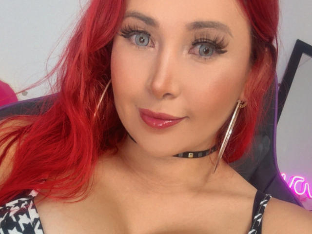 Close-up of NathaPeach with vibrant red hair, blue eyes, and full lips. She's wearing a black choker and large gold hoop earrings. Her tank top has a black-and-white pattern. Fucking sexy.