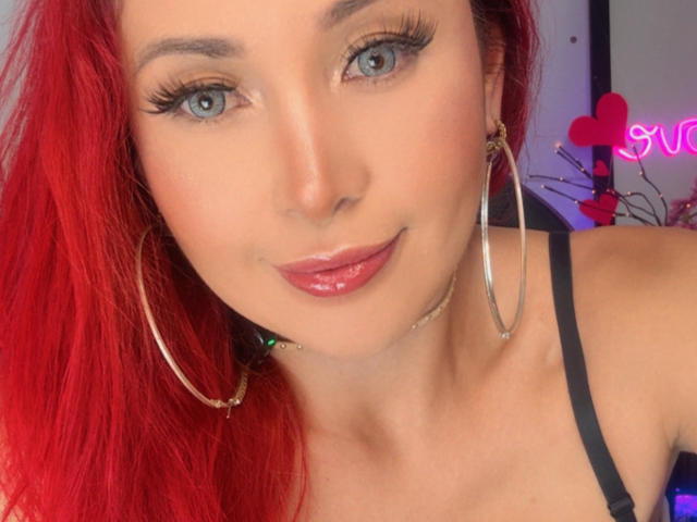 Close-up of NathaPeach with red hair, blue eyes, and glossy pink lips. She wears large hoop earrings and a black strap top. Neon pink hearts and "Love" sign in background. Fucking sexy.