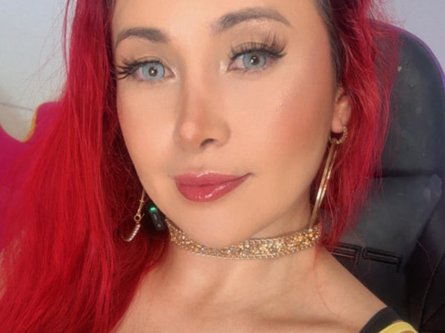 Close-up of NathaPeach with red hair, blue eyes, and glossy pink lips. She wears gold choker and earrings, looking directly at the camera with a slight smirk. Fucking hot.