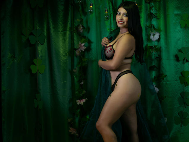 YinelaOslenX, mit üppigen Formen, großen Brüsten und einem runden Hintern, trägt schwarze Spitzendessous und steht vor einem dunkelgrünen Hintergrund. Vorhang mit Blumenmuster, Blick zurück mit sinnlicher Miene, langes schwarzes Haar, sexy Pose..