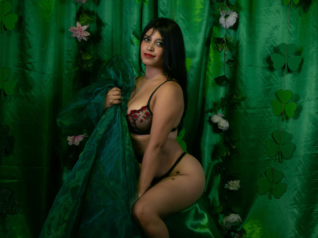 YinelaOslenX ajoelha-se sobre cortinas de seda verde, segurando um tecido verde translúcido. Ela veste lingerie de renda preta, com detalhes em vermelho no sutiã. Cabelo escuro, batom vermelho. Decorações com borboletas. Iluminação verde escura e sedutora..