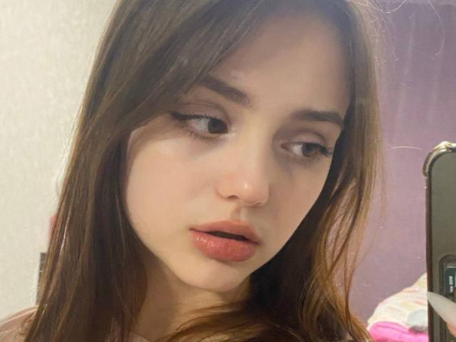 Primer plano de TrevaGiumarro con piel pálida, cabello castaño y labios rojos, mirando ligeramente hacia un lado con una expresión sensual. expresión, sosteniendo un teléfono inteligente en su mano derecha. El fondo está desenfocado..
