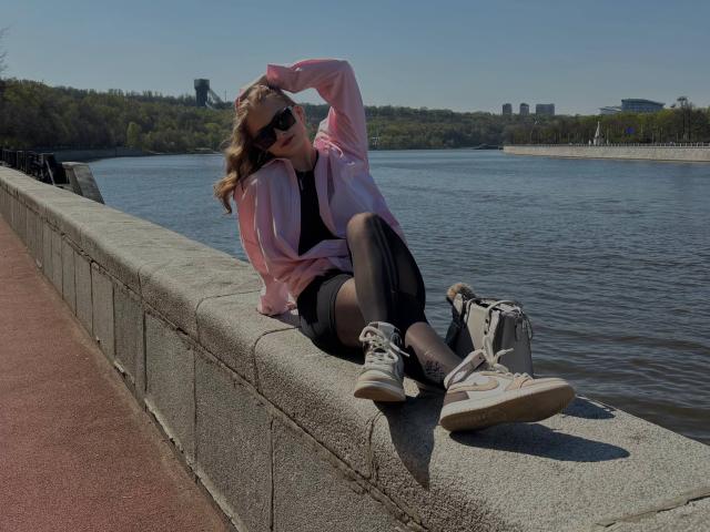 Maragret Gussman está sentada em uma parede de concreto perto de um lago, vestindo uma jaqueta rosa, uma saia preta e tênis brancos. Ela está recostada, com a mão no cabelo, transmitindo uma forte tensão sexual. Uma paisagem urbana aparece ao fundo..
