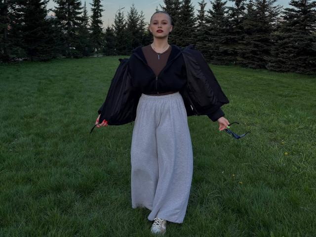 MaragretGussman standing in a grassy field, wearing a black top with puffed sleeves and loose, sparkly silver pants, holding a red object in each hand. Foreground and trees in background.