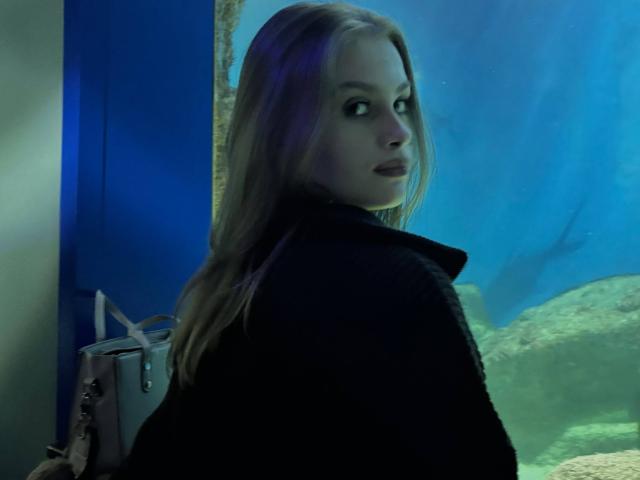 MaragretGussman with long blonde hair, turned back, looking over her shoulder, wearing a black coat, in a blue-lit setting with an aquarium background, intense gaze, sexual tension.