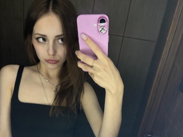 TrevaGiumarro si scatta un selfie allo specchio, tenendo in mano un telefono rosa con una fotocamera a forma di X nera. Ha la pelle chiara, i capelli castano scuro e indossa una canotta nera. Uno sguardo incredibilmente intenso..