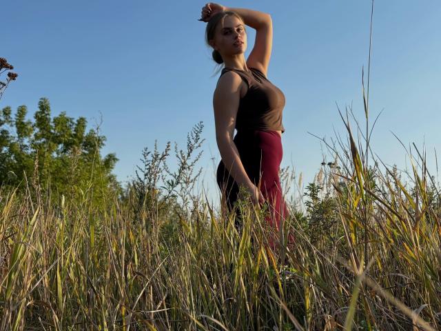 MaragretGussman stands in a tall grass field, wearing a black tank top and red pants, arm raised, sunlight casting shadows, clear blue sky, natural light, slight shadow on her face.