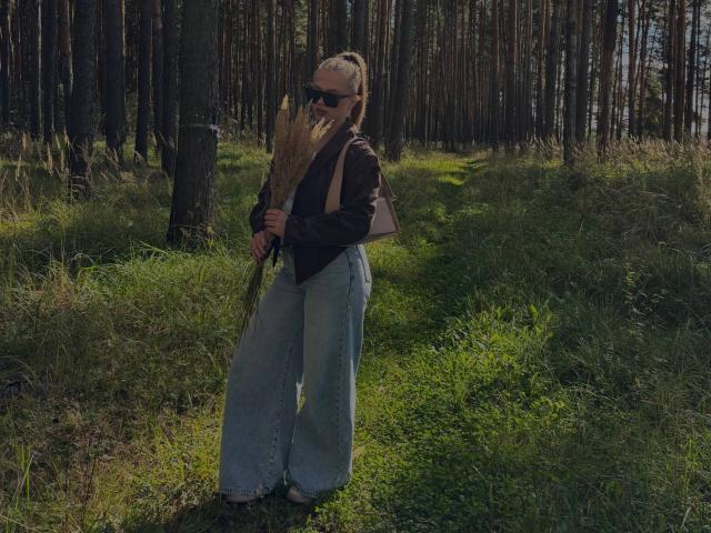 Maragret Gussman steht in einem Wald, trägt eine schwarze Jacke, blaue Jeans mit hoher Taille und eine Sonnenbrille und hält einen Stock. Hohe Bäume, die Schatten werfen, und grünes Gras umgeben sie. Sonnenlicht scheint hindurch. Verdammt heiß.