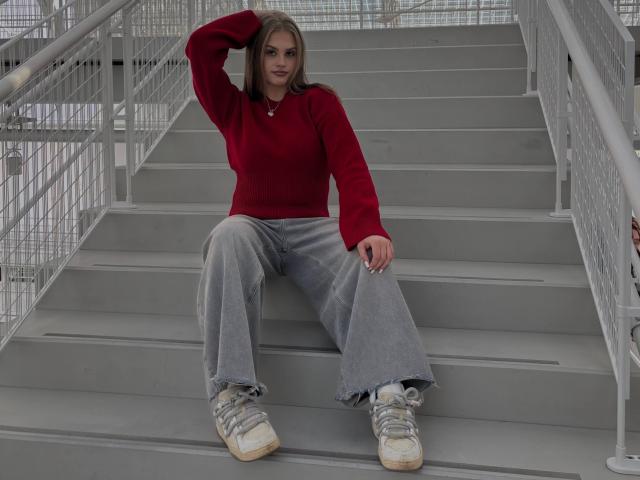 Maragret Gussman, com cabelo castanho liso, está sentada em degraus de metal cinza, vestindo um suéter vermelho, calças cinzas largas e brancas. tênis, mão no cabelo, olhar intenso, casual, ambiente urbano..