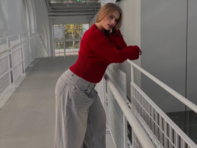 MaragretGussman leaning against a railing, wearing a red sweater and loose gray pants, her curvy hips and large ass emphasized. She's looking over her shoulder, blonde hair falling. Urban setting.