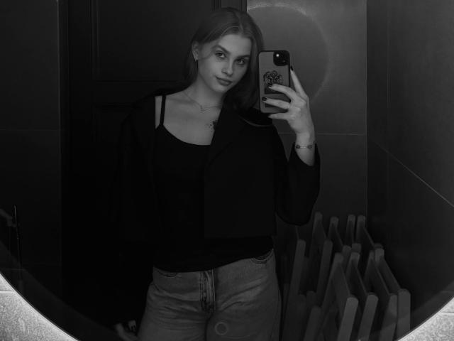 Monochrome photograph of MaragretGussman, standing in front of a mirror, holding a phone with a reflective case. She wears a black top and jeans, looking at the camera with a seductive expression.