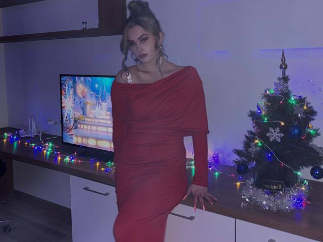Maragret Gussman está sentada em uma bancada, vestindo um vestido vermelho justo com um ombro de fora, com as pernas cruzadas e inclinada para trás. Uma árvore de Natal com luzes coloridas está à sua direita, com a tela da TV atrás..