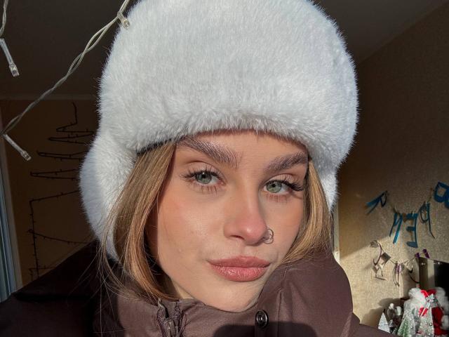 Close-up de Maragret Gussman, com pele clara, olhos verdes e um piercing no nariz, usando um chapéu de pele branca e um casaco marrom. A luz do sol realça seu rosto, criando sombras. Que fofa, uma expressão sexy..