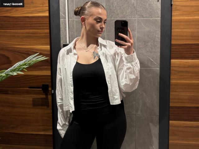 Maragret Gussman, riflessa nello specchio, tiene in mano un telefono, indossa una camicia bianca sopra una canotta nera e pantaloni, parete di legno. Sfondo: parete con piastrelle grigie, pannello in legno marrone scuro, pianta verde a sinistra. Assolutamente attraente..