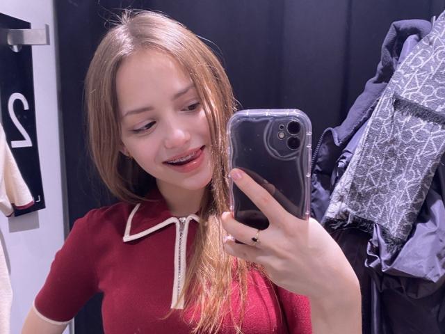 TrevaGiumarro si scatta un selfie allo specchio con un telefono nero, indossando una polo rossa con bordi bianchi. Lei sta sorridendo, con i capelli castano chiaro sciolti, e sullo sfondo si vede un appendiabiti con camicie a fantasia..